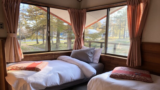 A bedroom with two beds, a large window with curtains, and a view of trees and a picnic table outside. - Alpha Lodge Madarao (Myoko)