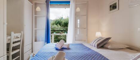A bedroom with a blue quilted bedspread, a white chair, a balcony with a table and chairs, and a window with curtains.
