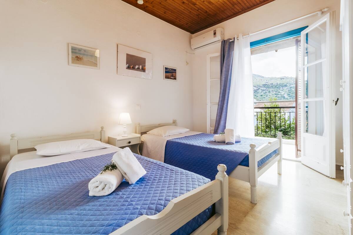 A bedroom with two beds, a nightstand, a lamp, and a view of the outdoors.