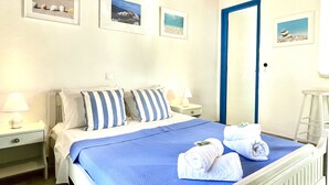 A neatly made bed with blue and white striped pillows, a blue bedspread, and two rolled towels on the bed. There are framed pictures on the wall, a nightstand with a lamp, and a blue door. - Sunflower Apartments & Studios (Corfu)