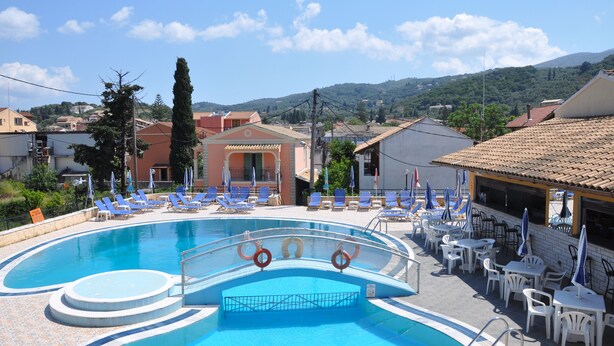 A swimming pool with sun loungers and umbrellas, surrounded by buildings and a view of hills. - Sunflower Apartments & Studios (Corfu)