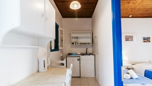 A compact hotel room with a kitchenette, a bed with blue bedding, a white table, and a white chair. - Sunflower Apartments & Studios (Corfu)