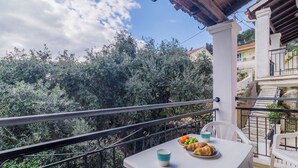A balcony with a table set for breakfast, including croissants and a bowl of fruit, overlooking a lush garden and a building with a red roof. - Sunflower Apartments & Studios (Corfu)
