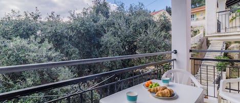 A balcony with a table set for breakfast, including croissants and a bowl of fruit, overlooking a lush garden and a building with a red roof.