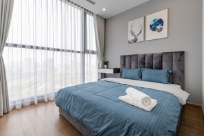 Un dormitorio moderno con una cama grande, un cabecero tapizado en gris y vistas de la ciudad a través de cortinas de gasa.