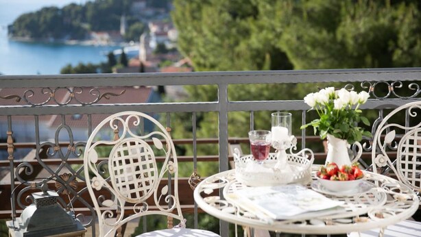A balcony with white metal chairs and a table set with a vase of flowers, a glass of drink, and a bowl of fruit.