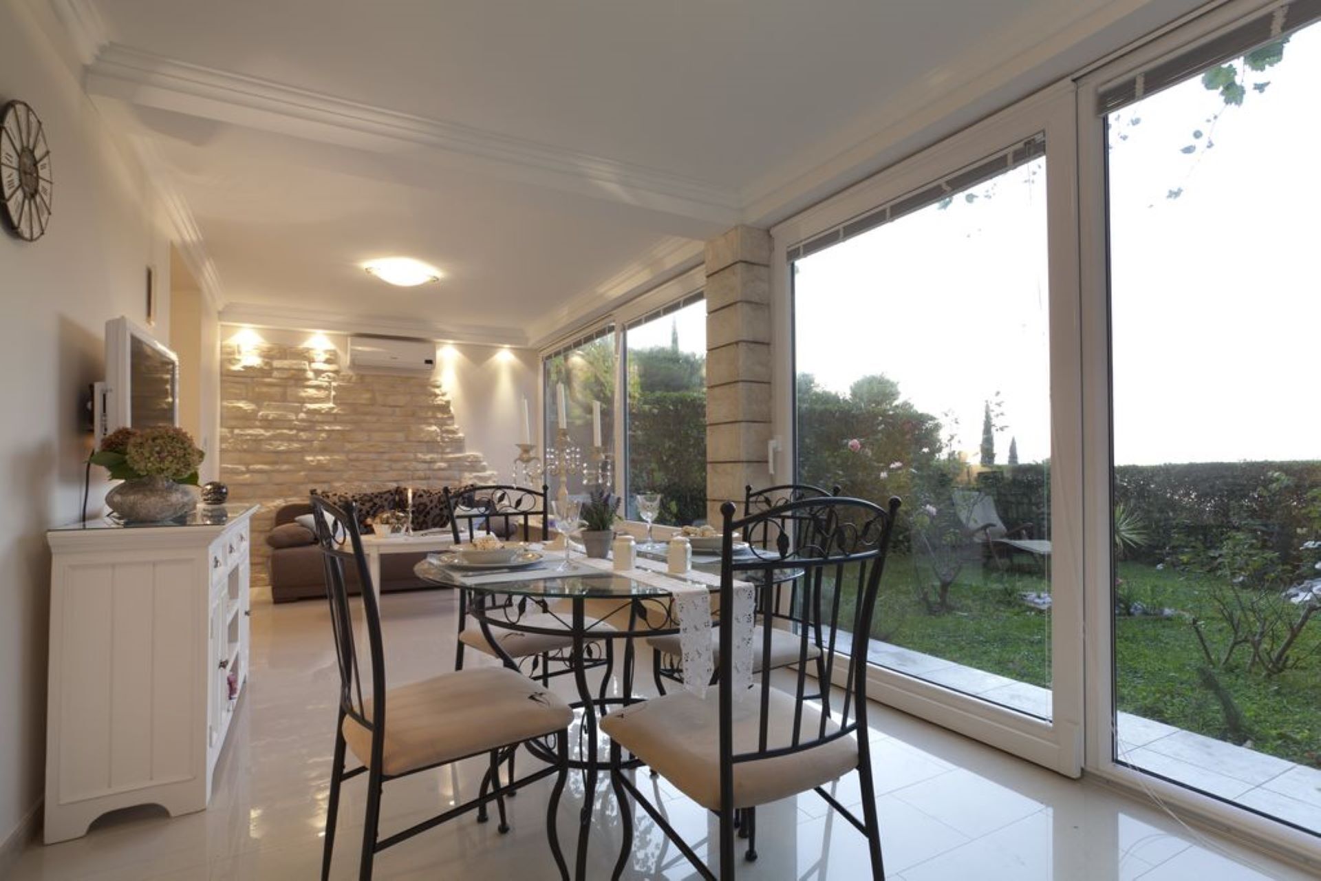 A modern kitchen with a dining area, large windows, and a stone wall.