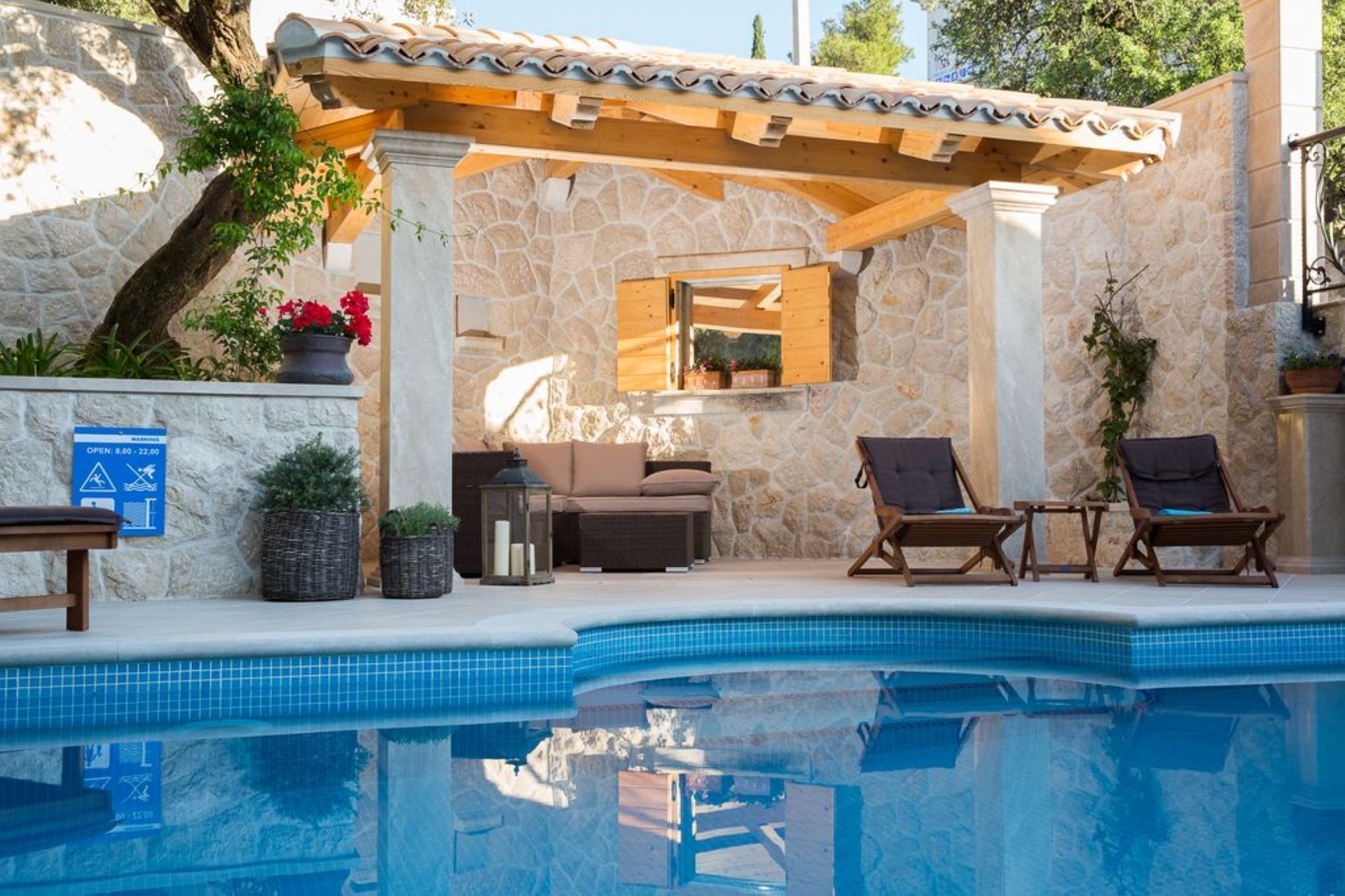 Ein Poolbereich mit Steinmauer, Holzpergola, Liegestühlen und einem kleinen Tisch.