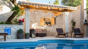 A pool area with a stone wall, wooden pergola, lounge chairs, and a small table.