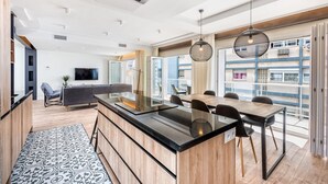 Una cocina moderna con una isla central, una zona de comedor con una mesa y sillas de madera, y ventanas amplias que ofrecen vistas de la ciudad.