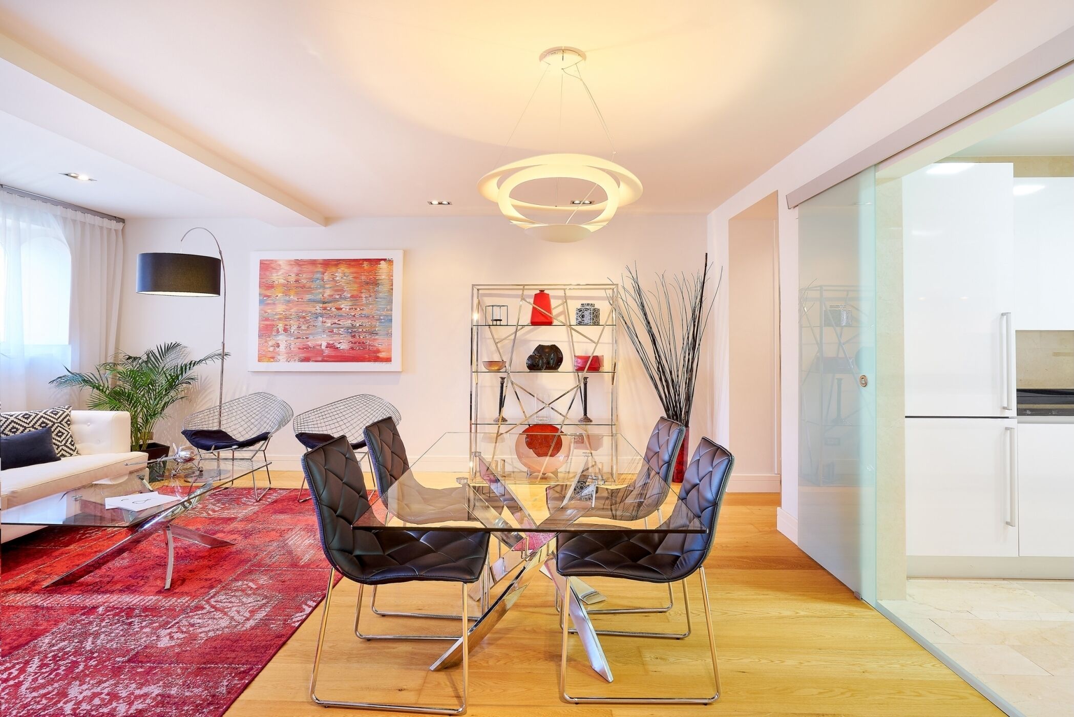 A modern living room with a glass dining table, black upholstered chairs, a red patterned rug, and a contemporary light fixture.