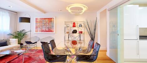 A modern living room with a glass dining table, black upholstered chairs, a red patterned rug, and a contemporary light fixture.