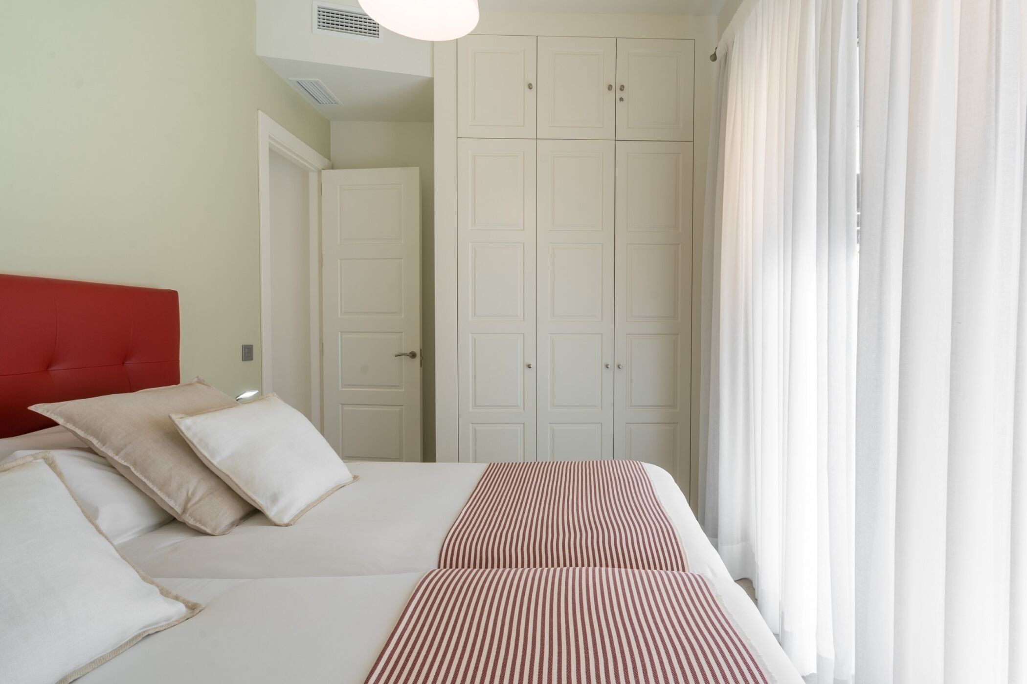 Un dormitorio con cabecera roja, cama con ropa de cama a rayas blancas y rojas, y armario blanco.