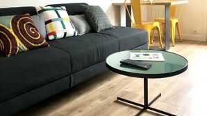 A modern living room with a black sofa, colorful pillows, a glass coffee table with a remote control, and a dining area with yellow chairs. - "L'Etale", apartment with terrace in the heart of the city center. 2 steps from the lake (Soorts-Hossegor)