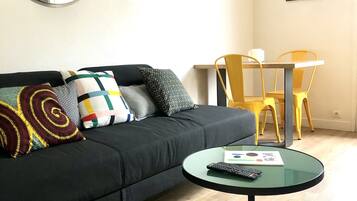 Un salon moderne avec un canapé noir, des coussins colorés, une table basse en verre avec une télécommande, et un coin repas avec des chaises jaunes.