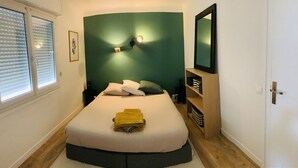 A bedroom with a bed, bedside tables, a nightstand, a wall-mounted lamp, a mirror, and a window with blinds. - "L'Etale", apartment with terrace in the heart of the city center. 2 steps from the lake (Soorts-Hossegor)