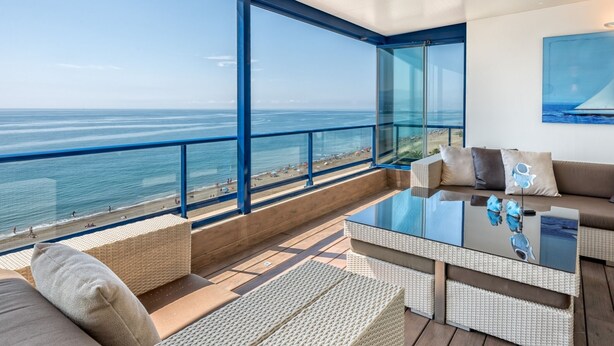 Un área de estar moderna al aire libre con una mesa de vidrio y mobiliario de mimbre con vista a una playa.