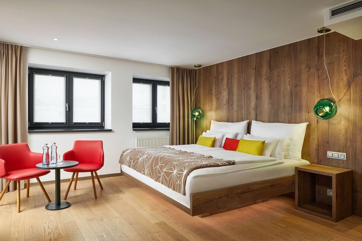 A modern hotel room with a large bed, a small table with chairs, and two windows with blinds.