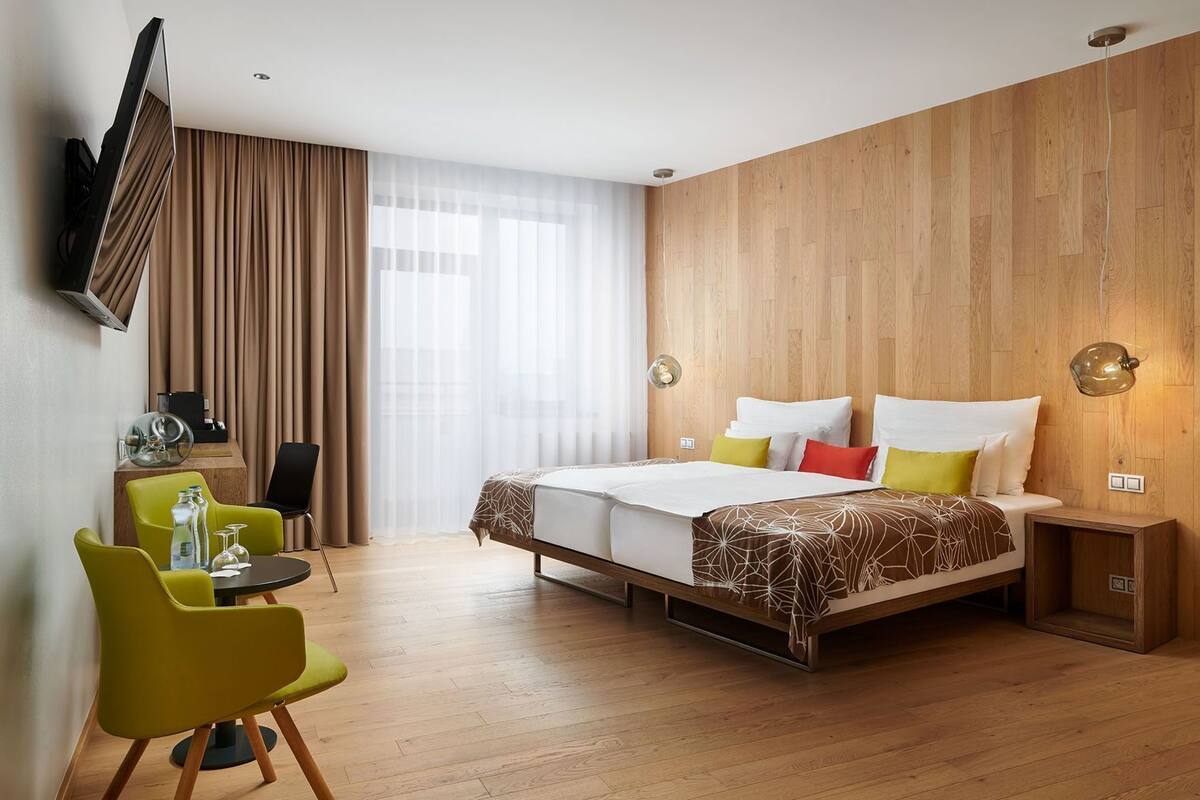 A hotel room with a bed, a desk with a chair, a TV mounted on the wall, and a wooden wall panel.