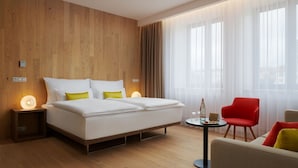 Una habitación de hotel moderna con una pared de paneles de madera, una cama grande con ropa blanca y almohadas amarillas, una silla roja, una pequeña mesa redonda con una botella y copas, y cortinas de encaje en las ventanas.