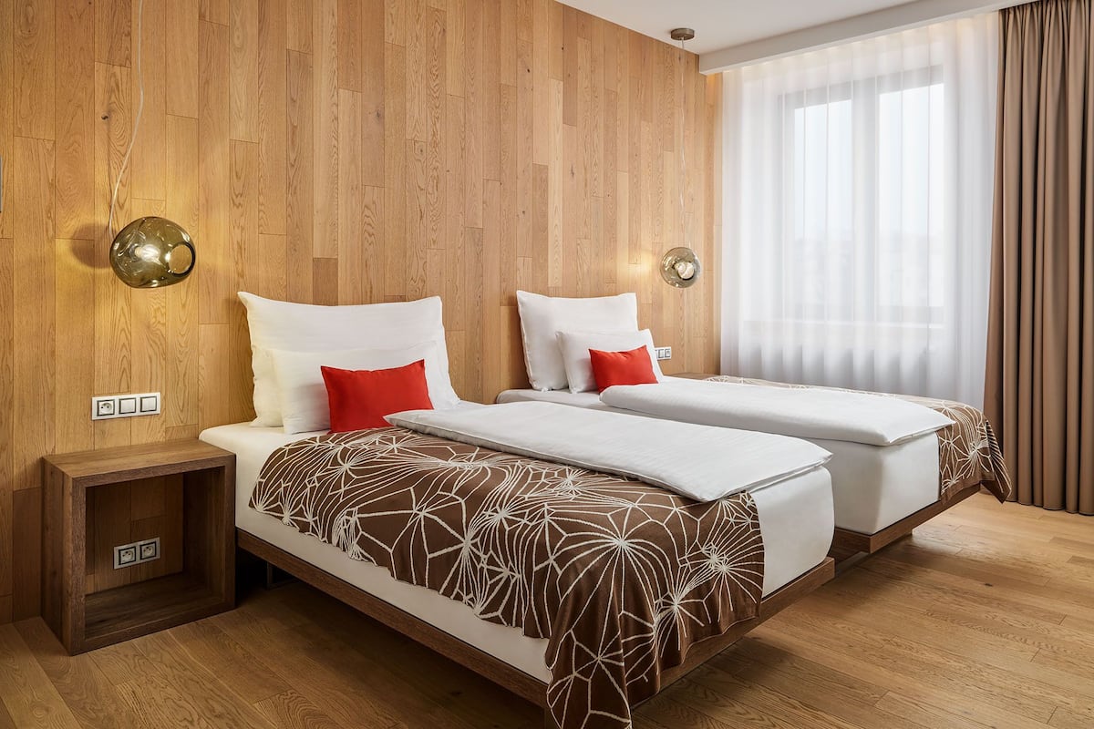 A double bed with a patterned bedspread, two bedside tables, and a large window with sheer curtains.