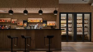Zona de bar con un bartender, mostrador de madera y taburetes. Se ven estantes con botellas al fondo.