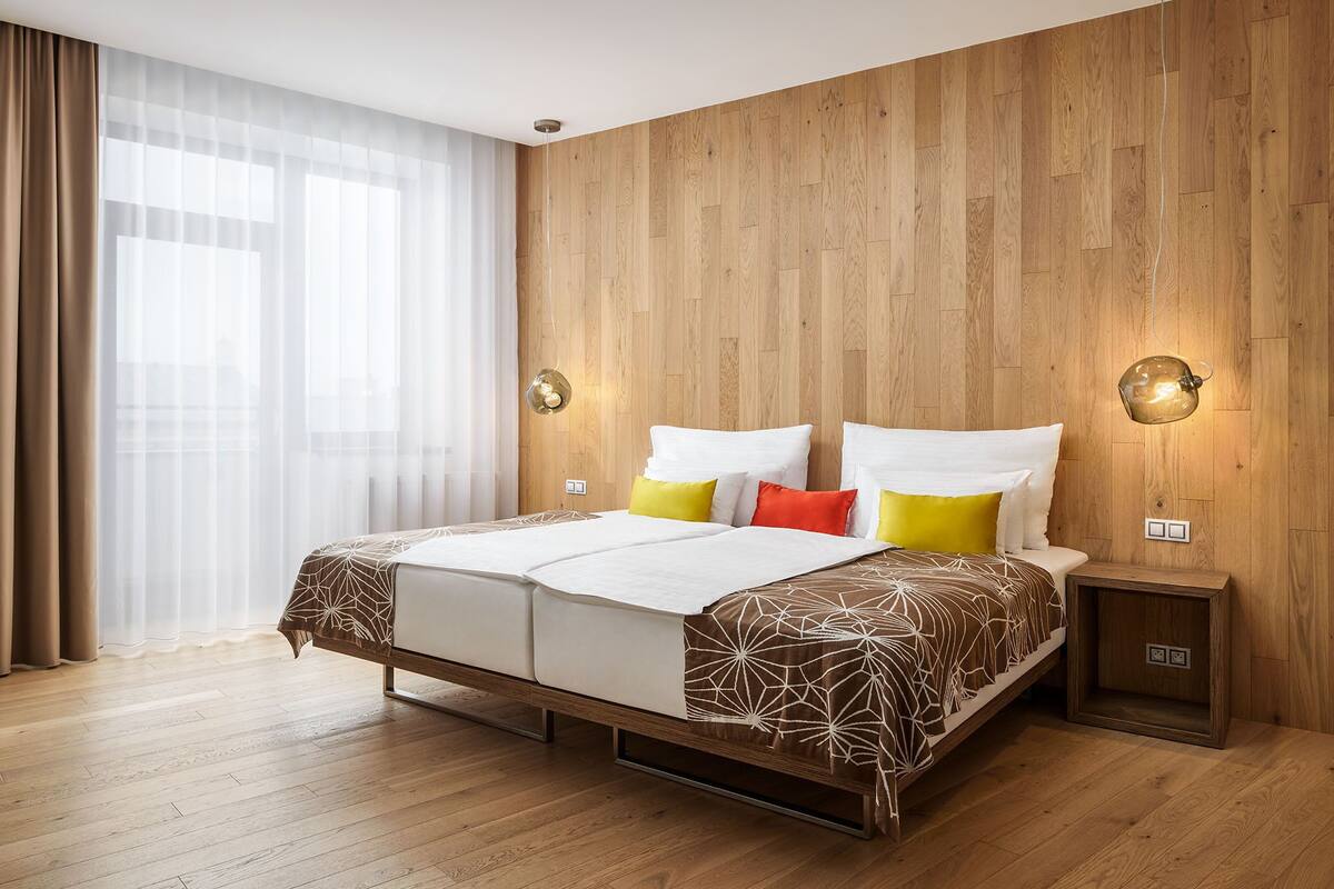 A modern hotel room with a wooden panel wall, a large bed with patterned bedding, and two wall-mounted light fixtures.