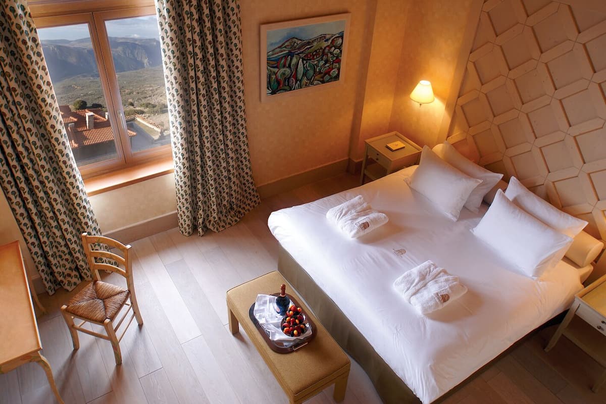 A hotel room with a bed, a chair, a bedside table with a lamp, a painting on the wall, and a window with curtains.
