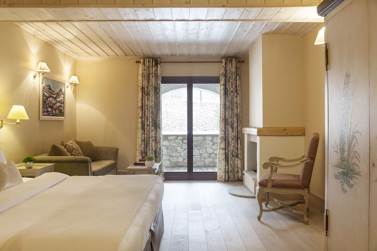 A hotel room with a bed, a sofa, a chair, a small table with a lamp, and a view through a glass door.