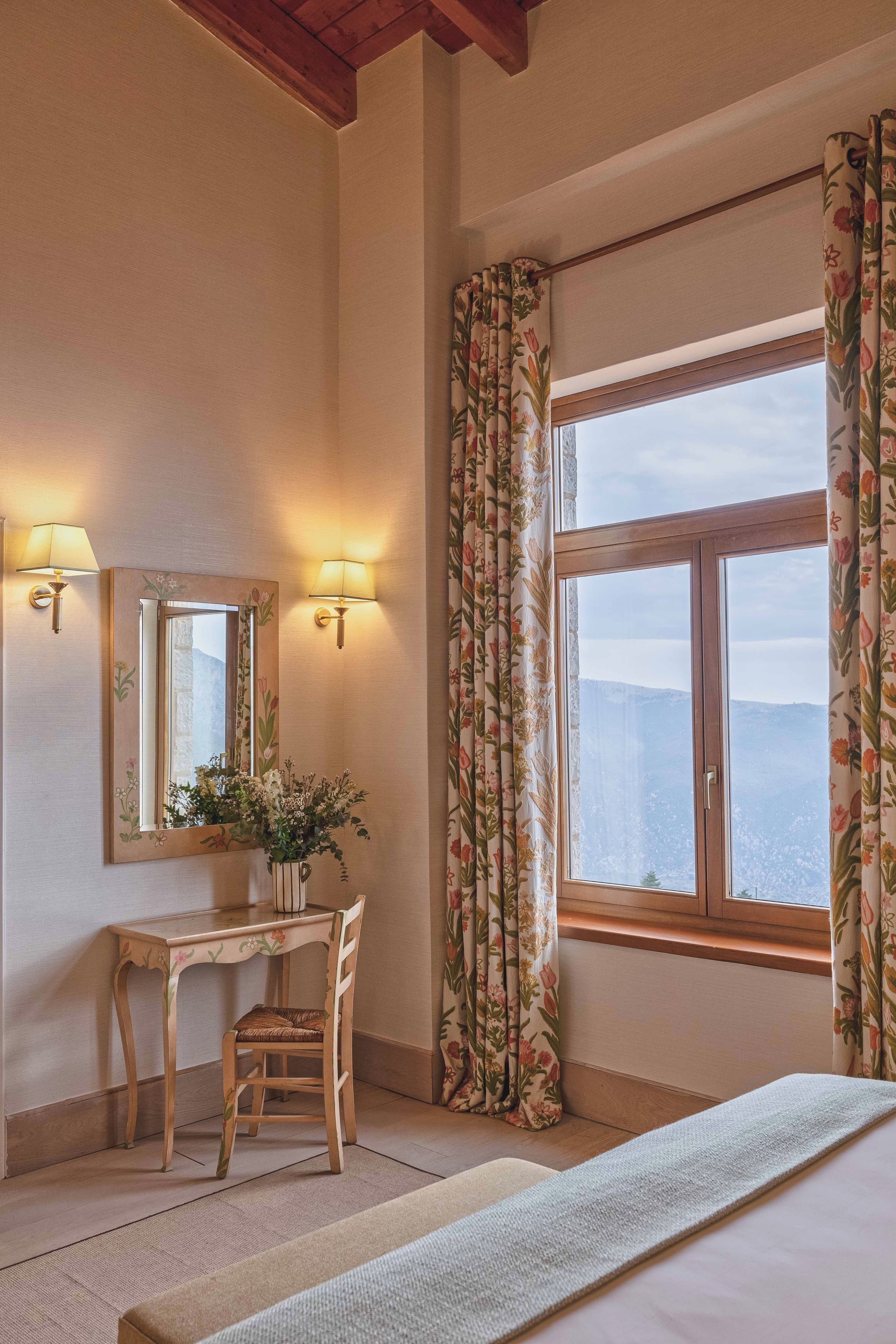 Habitación de hotel con escritorio de madera, una silla, un espejo y un ventanal con cortinas.