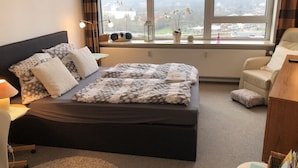 Ein modernes Hotelzimmer mit einem großen Bett, einer Sitzecke und Blick auf eine Stadtlandschaft durch große Fenster.