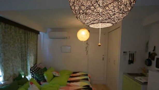 Quarto com cama, almofadas verdes, manta estampada, luminária pendente, pequeno espaço de cozinha e janela com cortinas.