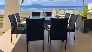 Uma área de estar coberta ao ar livre, com mesa e cadeiras, com vista para uma praia e oceano.