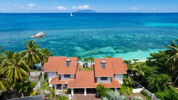 Uma casa de dois andares com telhado de telhas vermelhas, cercada por vegetação exuberante e vista clara para o oceano.