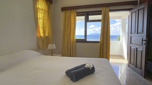 Quarto de hotel com uma cama, mesa de cabeceira, abajur e vista para o mar através de grandes janelas.