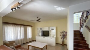 Uma sala de estar espaçosa com um sofá de couro marrom, uma mesa de centro branca, uma TV sobre um armário branco e uma escada com degraus de madeira.