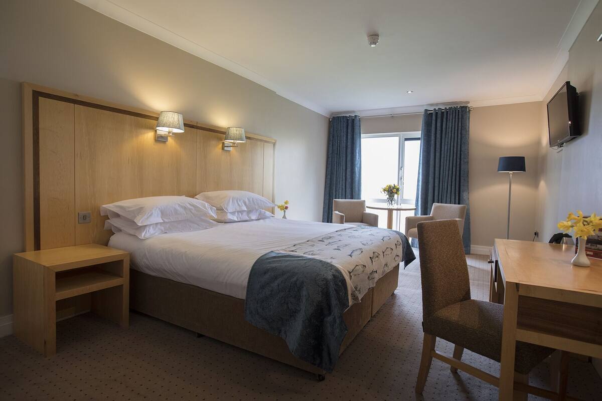 A hotel room with a large bed, bedside tables, a desk, a chair, a TV, and a window with curtains.