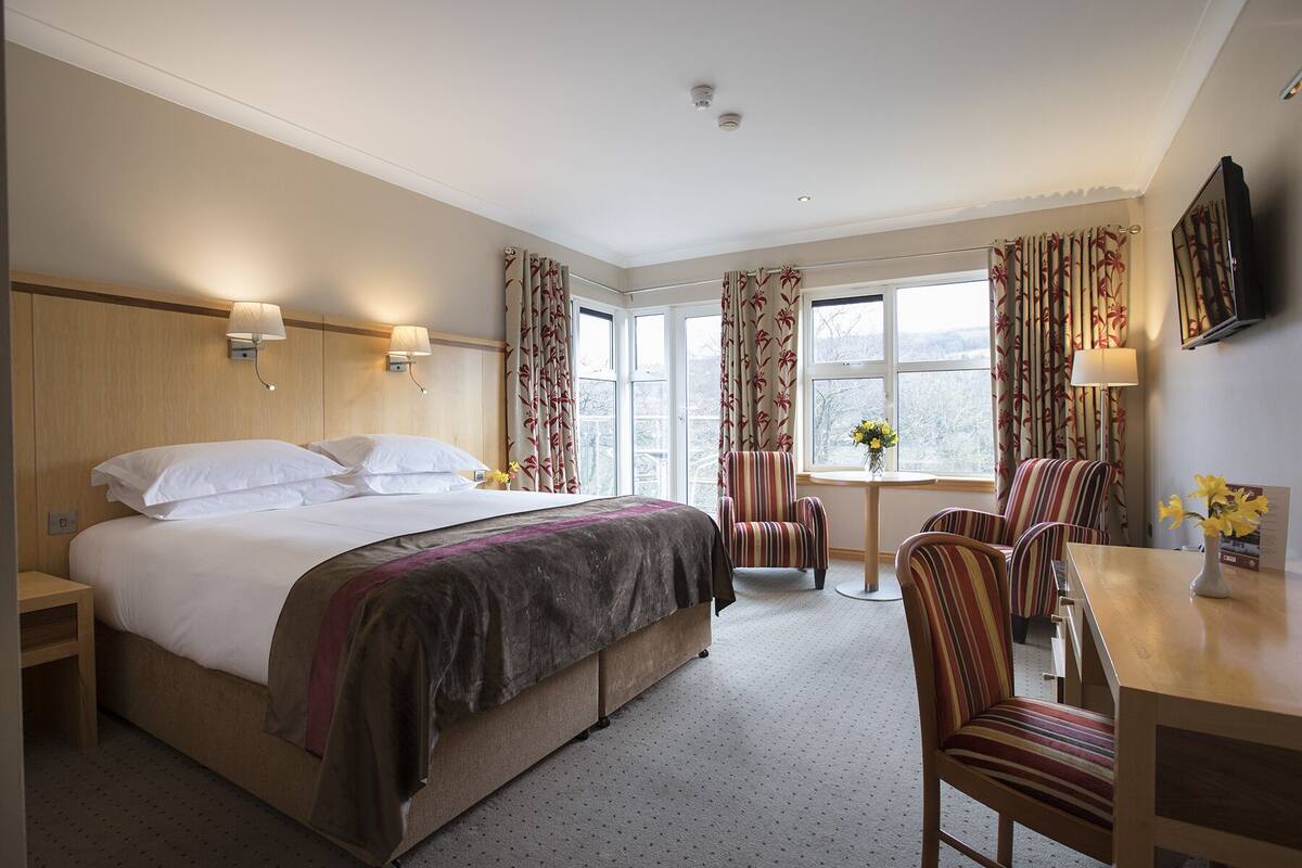 A hotel room with a large bed, a desk, two chairs, a small table, and a window with curtains.