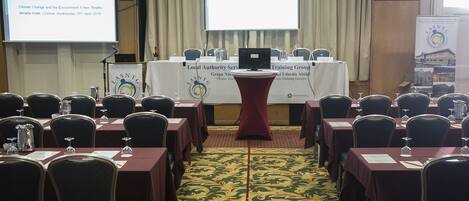 A conference room with tables and chairs arranged for a meeting. A projector screen and a podium are visible in the background.