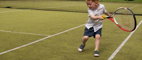 A child playing tennis on a court with a net, surrounded by trees and a cloudy sky.