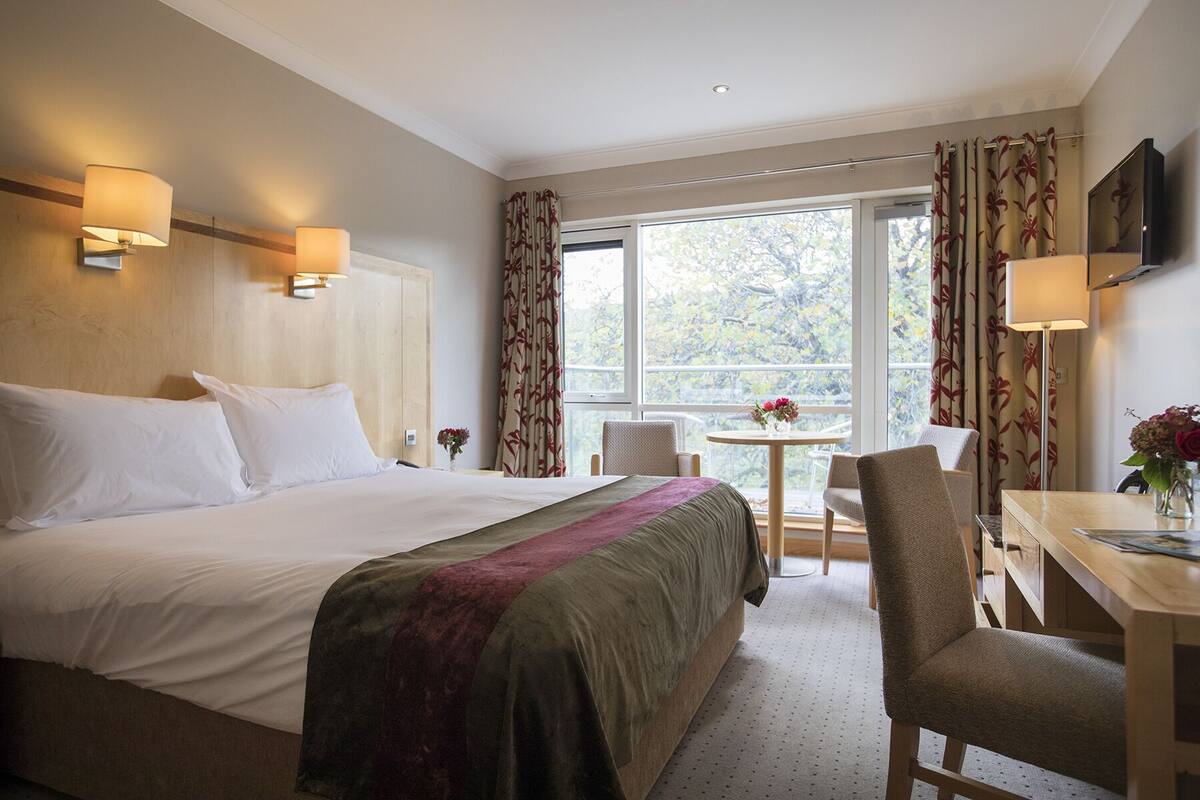 A hotel room with a large bed, a desk, and a chair. There is a window with curtains, a small table with a vase of flowers, and a television mounted on the wall.