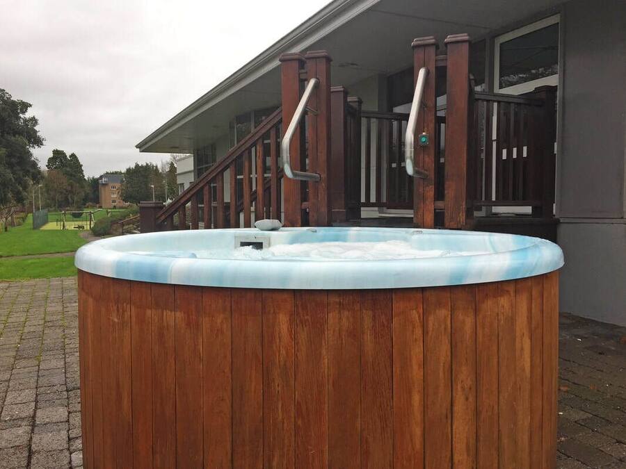 A wooden hot tub with metal handrails in front of a building with large windows.