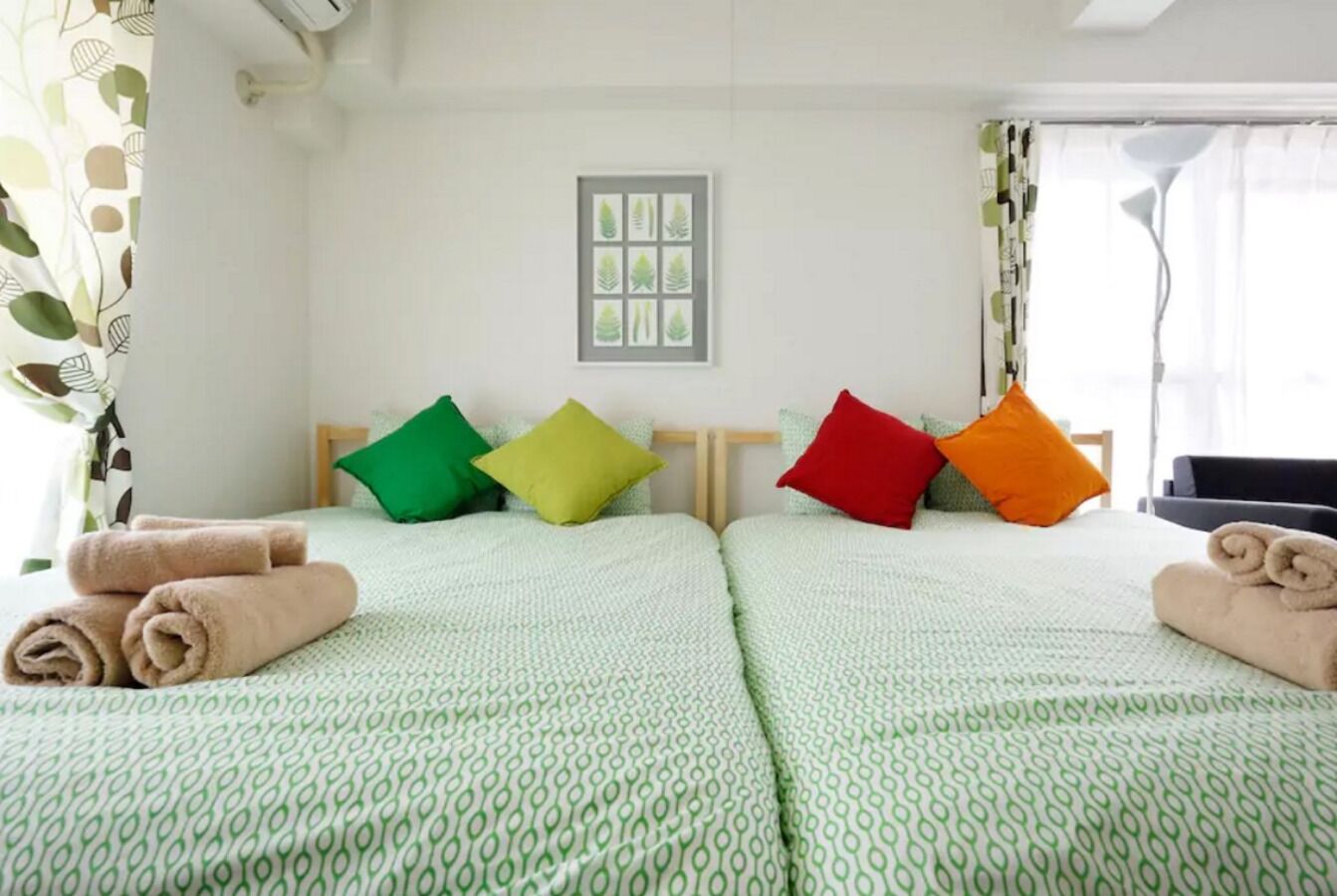 Ein Hotelzimmer mit zwei Betten, jeweils mit einer grün gemusterten Tagesdecke, einem gerahmten Bild an der Wand und bunten Kissen.