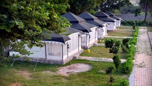 Une rangée de tentes blanches, surmontées de toits noirs, est disposée dans un terrain de camping aménagé avec un chemin pavé et de petits arbres.