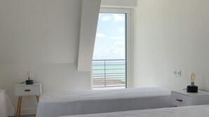 A modern bedroom with two beds, a nightstand, and a view of the sea through a large window. - Villa FEUNTEUN on the ocean! (Clohars-Carnoët)