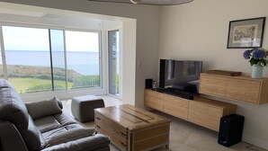 A modern living room with a large window offering a view of the sea, a grey sofa, a wooden coffee table, and a TV on a wooden cabinet. - Villa FEUNTEUN on the ocean! (Clohars-Carnoët)