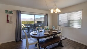 A dining area with a dark wooden table, white chairs, and a bench. There is a view through the window, a chandelier, and a sign on the wall that says "Eat". - Las Palmas Resort 704, Newly Renovated Main level 3 bd unit, Close to pool + More (St. George)