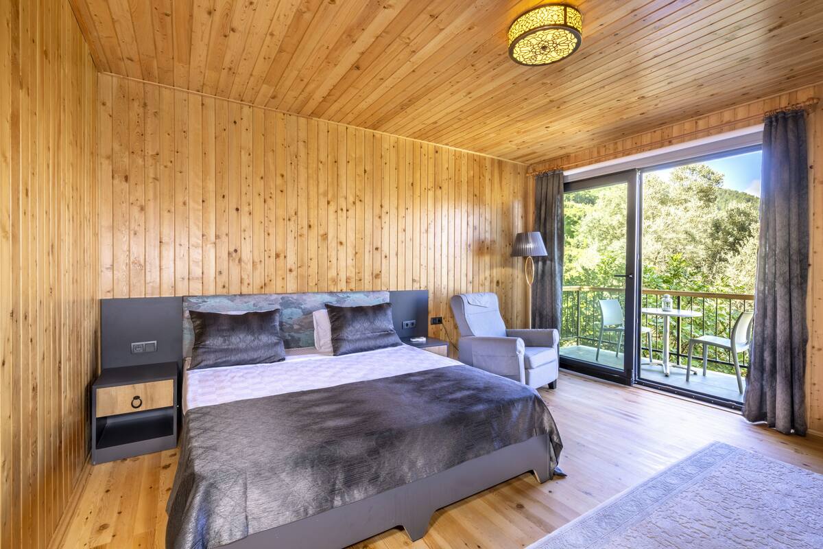 Un dormitorio con paneles de madera, una cama grande, una mesita de noche, una silla y un balcón con vistas a zonas verdes.
