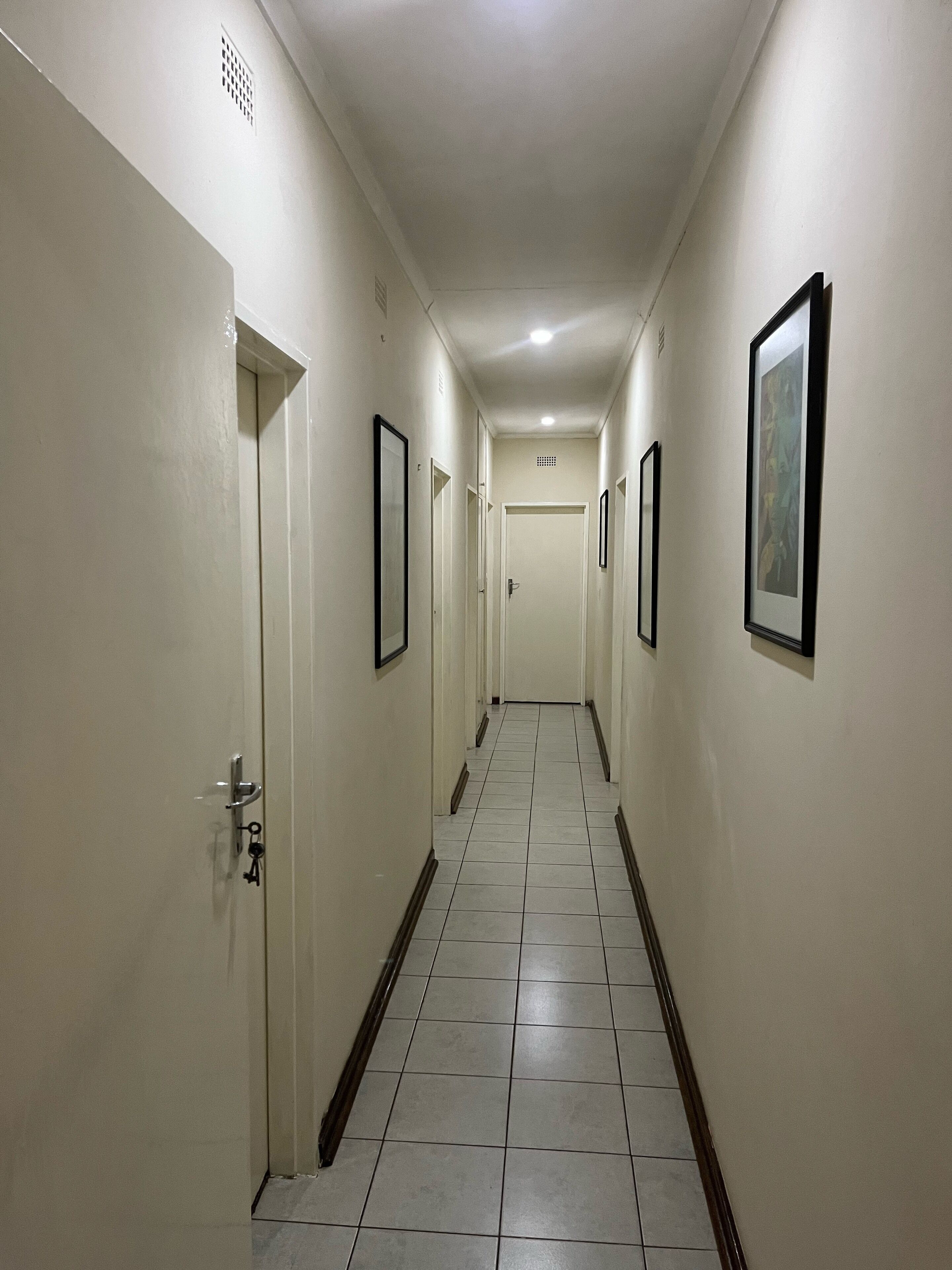 A long, narrow hallway with white walls and tiled flooring, featuring doors and framed pictures on the walls.