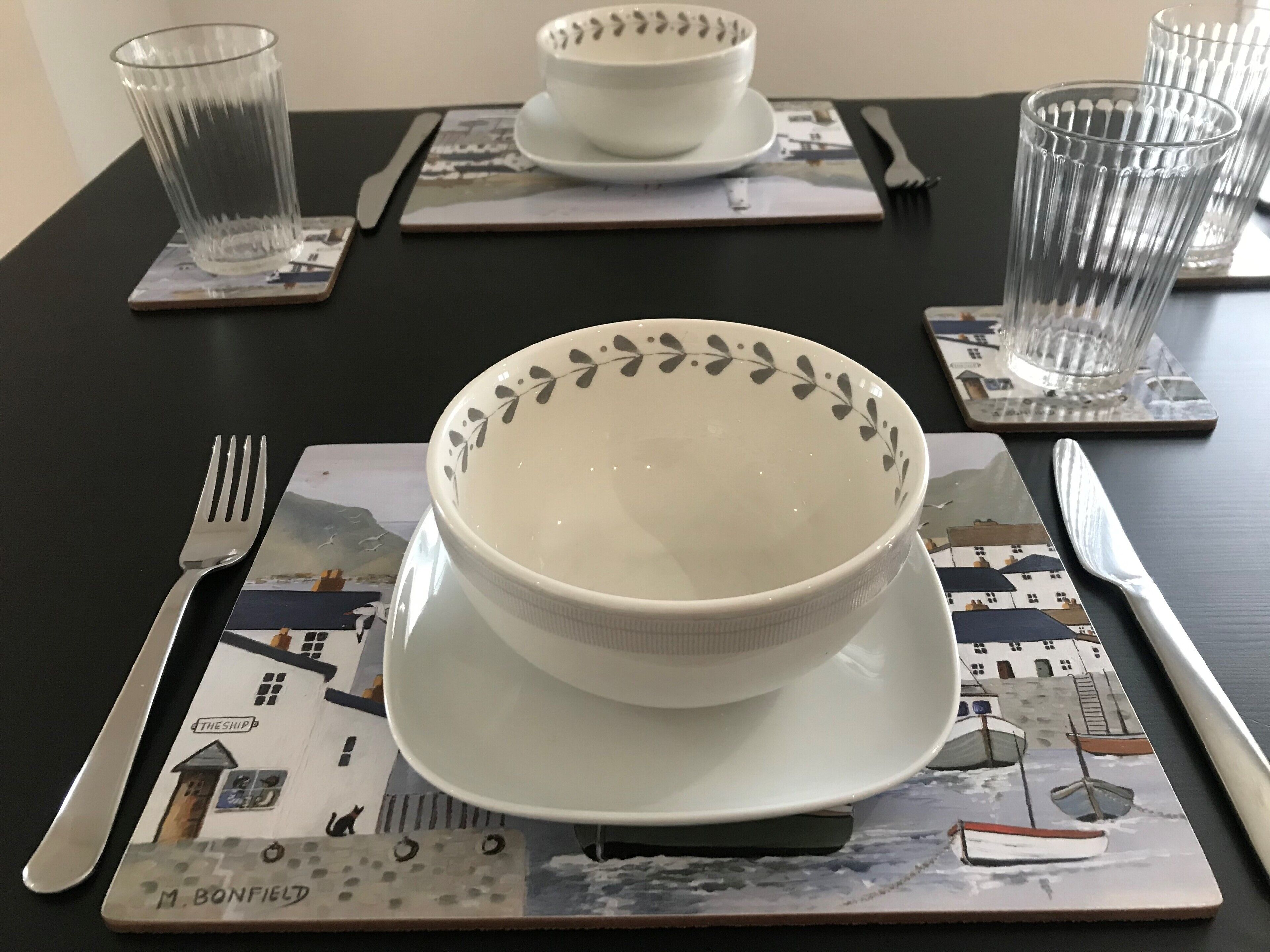A dining table set with a white bowl, a fork, a knife, and a glass on a placemat with a nautical theme.